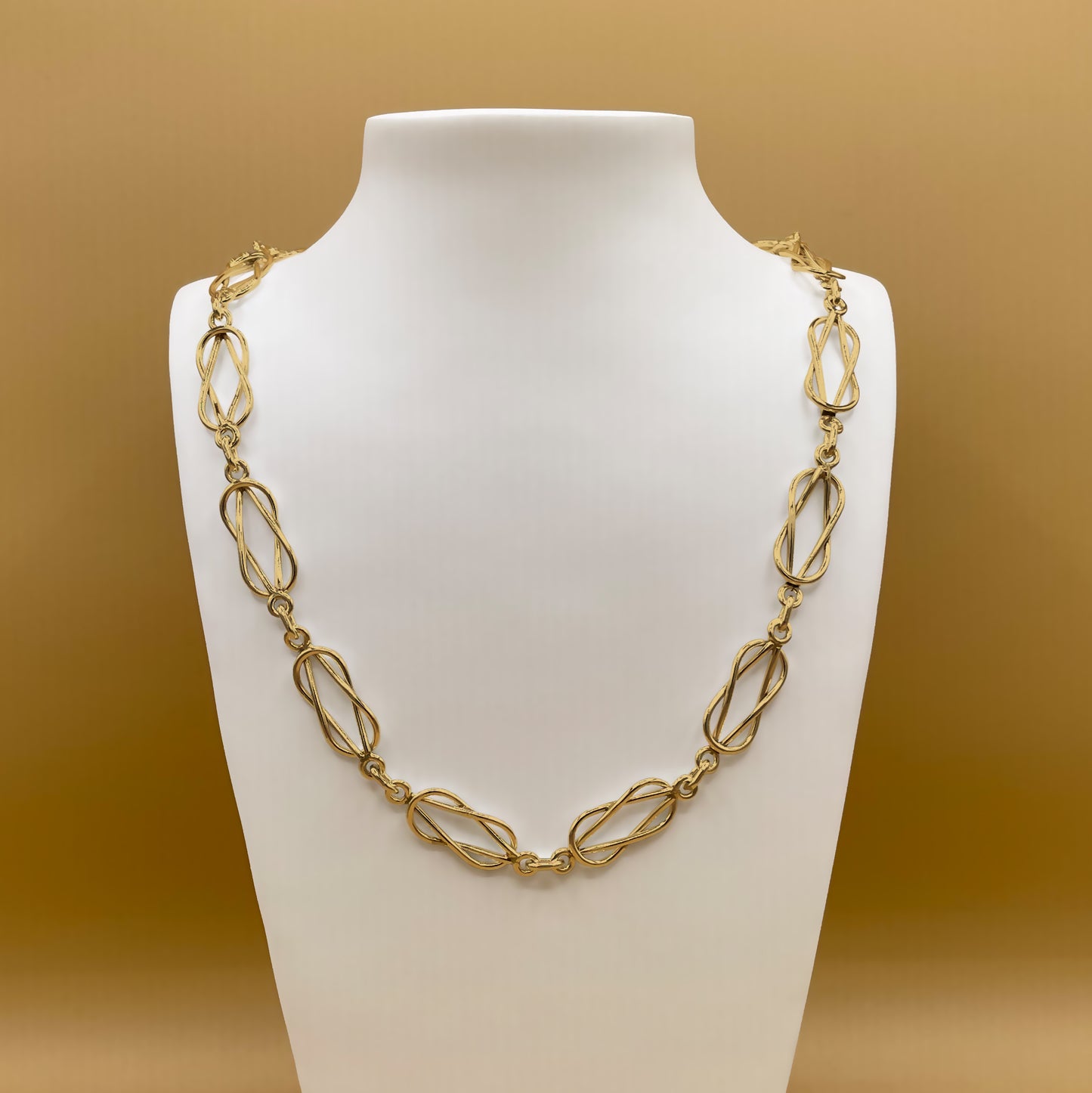 Timio's Forget-Me-Knot chain, in 24 karat gold, displayed on a white bust in front of a golden background.