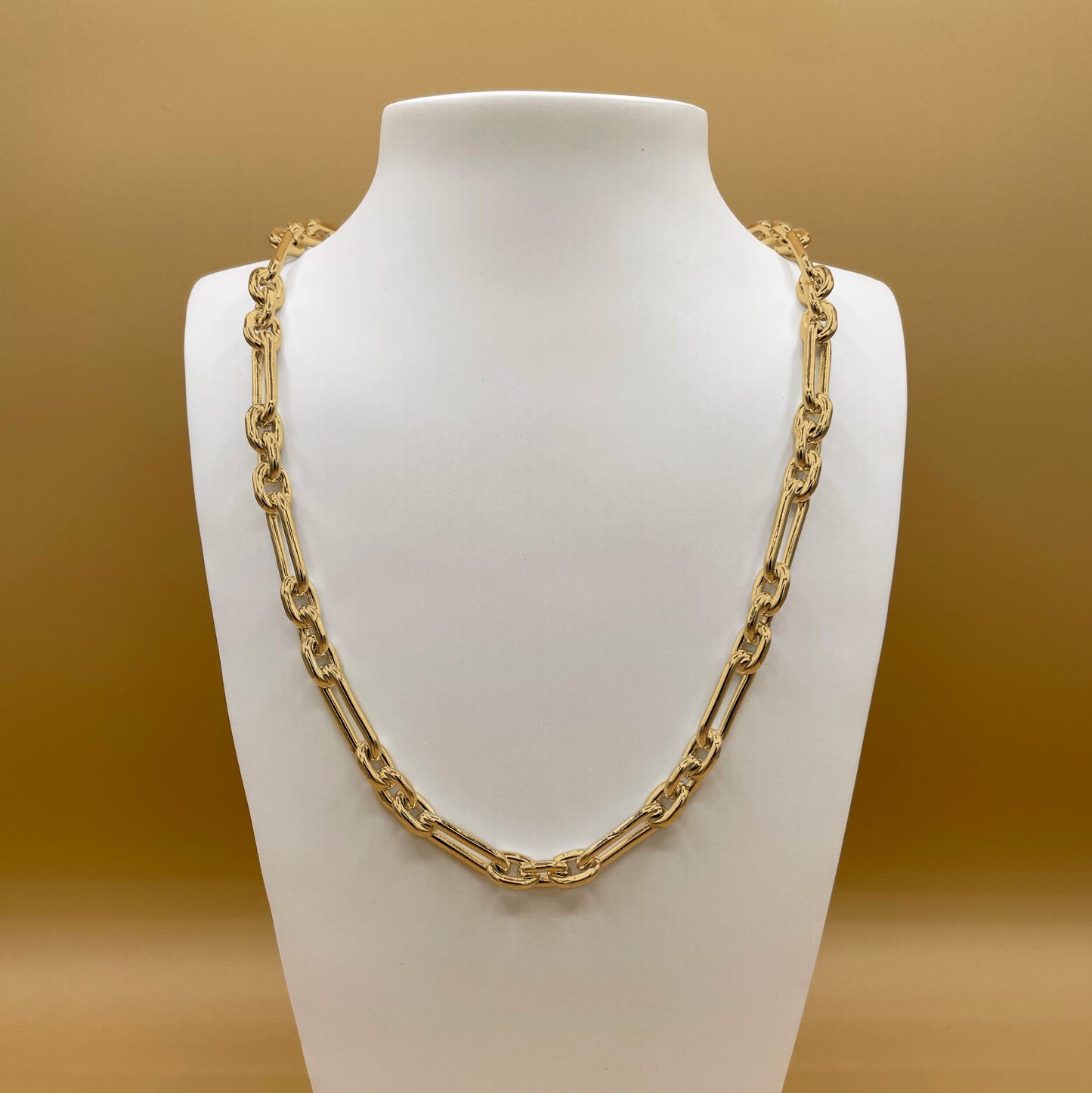 The Makarios style chain in 24 karat gold on a white bust stand in front of a light, golden background.